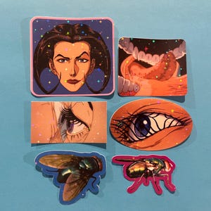 May include: A collection of five stickers with various illustrations. One sticker features a woman's face with dark hair, another depicts a mouth with a tongue, and two show eyes. The last two stickers are of flies.