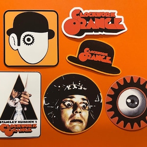 May include: A selection of stickers with designs from the film "A Clockwork Orange." The stickers display the film's title, a bowler hat, a character's face, and an eye with a gear design. The stickers are in orange, black, white, and red.