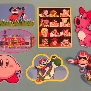 May include: A collection of colorful stickers featuring characters from classic video games. Includes a red dog with a duck, characters from Street Fighter, a pink dinosaur, Kirby, Mario and Yoshi, and a surfing monkey.