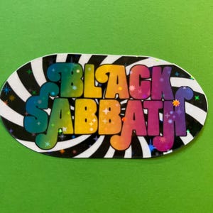 May include: Oval sticker with the text "BLACK SABBATH" in colorful, bubble-style letters. The letters transition from blue to purple, with yellow and orange accents. The background is a black and white striped pattern on a green background.