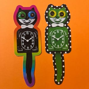 May include: Two cat-shaped clocks with whimsical designs. One is black with a rainbow tail and the other is green with a polka-dot border. Both have white bow ties and clock faces with black numbers and hands.