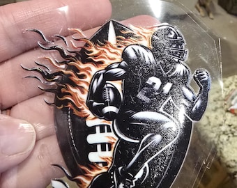 May include: A black and white football player graphic with flames. The player is wearing a helmet and holding a football. The graphic is on a clear, plastic-like material.
