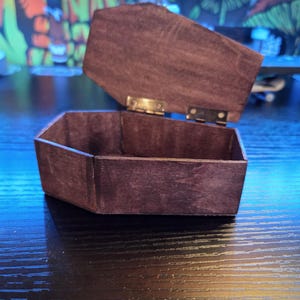 Könnte beinhalten: Eine offene, sechseckige Holzbox mit einem Klappdeckel. Die Box ist dunkelbraun gebeizt, die Scharniere sind goldfarben. Die Box steht auf einer dunklen, reflektierenden Oberfläche.