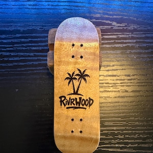 Pode incluir: Um fingerboard de madeira com acabamento castanho claro e topo prateado. A prancha apresenta um gráfico de palmeira e o texto "RIVRWOOD" em preto. Quatro furos são perfurados para os trucks. O fingerboard está apoiado num bloco de madeira.