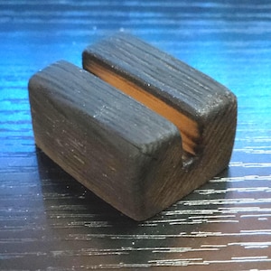 May include: A small, dark-stained wooden phone stand with a slot for holding a phone. The stand is rectangular with a groove cut into the top. The text "Rivrwood Fingerboards" is at the bottom.