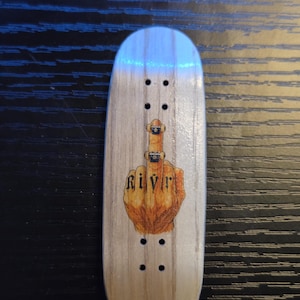 Könnte beinhalten: Ein Fingerboard-Deck aus Holz mit einer Grafik einer Hand, die den Mittelfinger zeigt, mit dem Wort "Rivr" darunter. Das Deck hat vier Sätze von zwei Löchern. Das Holz hat eine helle, natürliche Farbe.