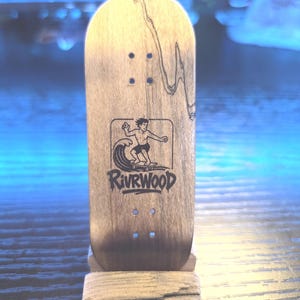 May include: A wooden fingerboard with a surfer graphic and the text "Rivrwood". The board is light brown with dark wood grain and has four small holes for the trucks. It is displayed on a small wooden stand.