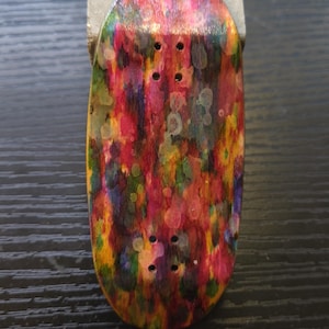 Peut inclure: Une planche de fingerboard présentant un motif abstrait aux éclaboussures de peinture multicolores vives, avec des nuances de rouge, jaune, vert et violet. La planche en bois comporte quatre paires de trous de montage pré-percés pour les trucks.