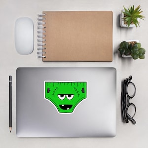 Könnte beinhalten: Ein grüner Aufkleber mit einem Cartoon-Design von Frankensteins Monster-Unterwäsche. Die Unterwäsche hat einen linealartigen Rand und genähte Details, mit einem lächelnden Gesicht und zwei Augen. Der Aufkleber befindet sich auf einem Laptop.