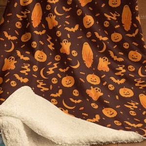 May include: A brown Halloween-themed throw blanket with an orange pattern of pumpkins, ghosts, bats, and crescent moons. The blanket has a soft, cream-colored sherpa lining, perfect for cozy autumn nights. The blanket is draped over a brown leather couch.