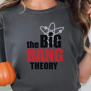 May include: Dark gray t-shirt with the text "the BIG BANG THEORY" in black and red, and a white atom symbol. The shirt is worn with blue jeans. An orange mug is held in the hand. A "BEST SELLING" sticker is in the top right corner.