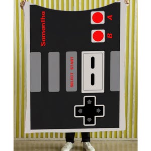 May include: A black blanket featuring a retro video game controller design. The blanket has red buttons, gray rectangles, and the text "Samantha" in red. The design is reminiscent of classic gaming consoles.