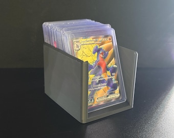 Può includere: Un porta carte grigio pieno di carte collezionabili. Le carte sono in custodie protettive, con una carta dorata e viola visibile. La carta presenta un'illustrazione di personaggio e testo.