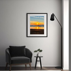 Könnte beinhalten: Ein gerahmter Druck eines Sonnenuntergangs über dem Meer. Der Himmel zeigt eine Mischung aus Orange, Gelb und Blau, wobei sich die Sonne im Wasser spiegelt. Der Druck hängt an einer weißen Wand, mit einem schwarzen Stuhl und einem Beistelltisch im Vordergrund.