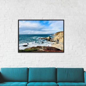 Könnte beinhalten: Gerahmtes Landschaftsfoto einer Küstenlandschaft. Das Bild zeigt das Meer mit Wellen, die gegen Felsklippen und einen Sandstrand schlagen. Der Himmel ist blau mit verstreuten Wolken, und der Vordergrund zeigt grüne und rote Vegetation.
