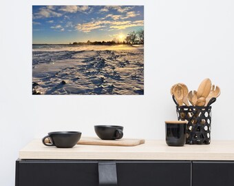 May include: A square print of a winter landscape with a snow-covered field under a cloudy blue sky. The sun sets on the horizon, casting a golden glow. Black cups and kitchen utensils are on a wooden counter.