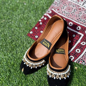 Puede incluir: Un par de bailarinas de terciopelo negro con intrincados adornos de perlas y cuentas doradas. Los zapatos tienen suelas de cuero marrón y un diseño bordado en oro en la puntera. Están sobre hierba verde, con un textil rojo y blanco estampado en el fondo.