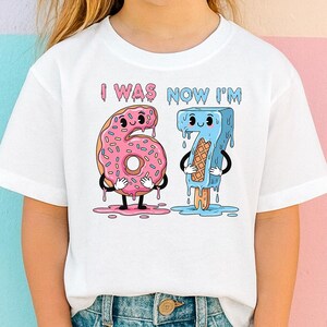 May include: White t-shirt with the text "I WAS NOW I'M" above a cartoon graphic of a pink donut and a blue popsicle. The donut is shaped like the number 6, and the popsicle is shaped like the number 7.