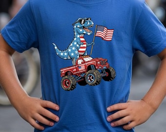 Puede incluir: Una camiseta azul real de manga corta con un estampado gráfico de un Tyrannosaurus Rex con gafas de sol y un patrón de estrellas, de pie sobre un camión monstruo rojo mientras sostiene una bandera estadounidense.