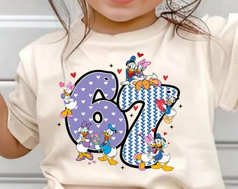 Puede incluir: Una camiseta color crema con el número "67" en un diseño morado y azul. El número está decorado con personajes de dibujos animados y pequeños corazones rojos. La camiseta la lleva un niño pequeño.