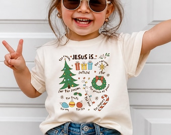 May include: A cream-colored short-sleeve t-shirt featuring a colorful Christmas-themed graphic. The design includes a green tree, presents, a wreath, a candy cane, and ornaments with text reading: The reason JESUS IS... faithful, the morning star, the prince of peace, the truth, the vine, the way, the life, the shepherd, and for the season.