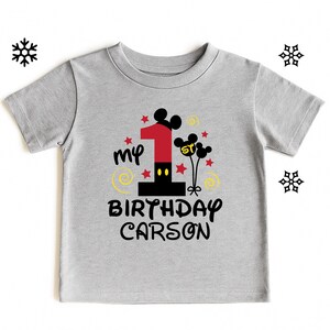 K&ouml;nnte beinhalten: Graues Kurzarm-T-Shirt mit farbenfrohem Grafikdesign f&uuml;r den ersten Geburtstag. Das Design umfasst eine gro&szlig;e rote Zahl 1, Mickey Mouse Ohren, Sterne und den Text "my BIRTHDAY CARSON."