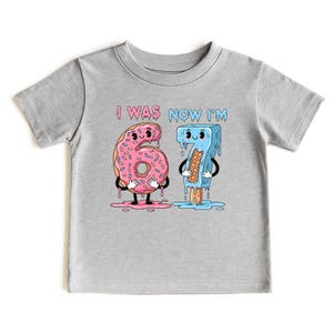 May include: A gray t-shirt featuring a cartoon graphic of a pink donut and a blue popsicle, each shaped like the numbers 6 and 7. The donut has sprinkles and the popsicle holds a waffle. The text "I WAS NOW I'M" is above the numbers.
