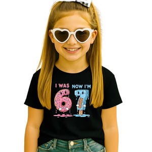 May include: A young girl wearing a black t-shirt with the text "I WAS NOW I'M" above a pink donut with the number 6 and a blue popsicle with the number 7. She is also wearing white heart-shaped sunglasses.