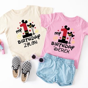 K&ouml;nnte beinhalten: Zwei Kinder-T-Shirts, eines cremefarbenes und eines rosa, mit einem Mickey Mouse "Mein 1. Geburtstag"-Design. Das cremefarbene Shirt tr&auml;gt die Aufschrift "Zylon" und das rosa Shirt "Derek". Accessoires sind karierte Schuhe und blaue Shorts.