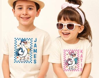 May include: Two off-white short-sleeve t-shirts featuring graphic prints of geese. One shirt displays a blue checkered border with a goose wearing a cap and backpack, labeled JAMES. The other has a pink checkered border with a goose wearing a bow and backpack, labeled KATIE.