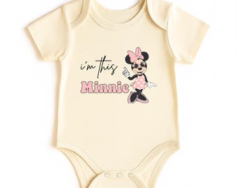 May include: Cream-colored baby bodysuit featuring a Minnie Mouse graphic. The graphic includes the text "I'm this Minnie" in black and pink. Minnie Mouse is depicted wearing a pink polka-dot dress, bow, and sunglasses.