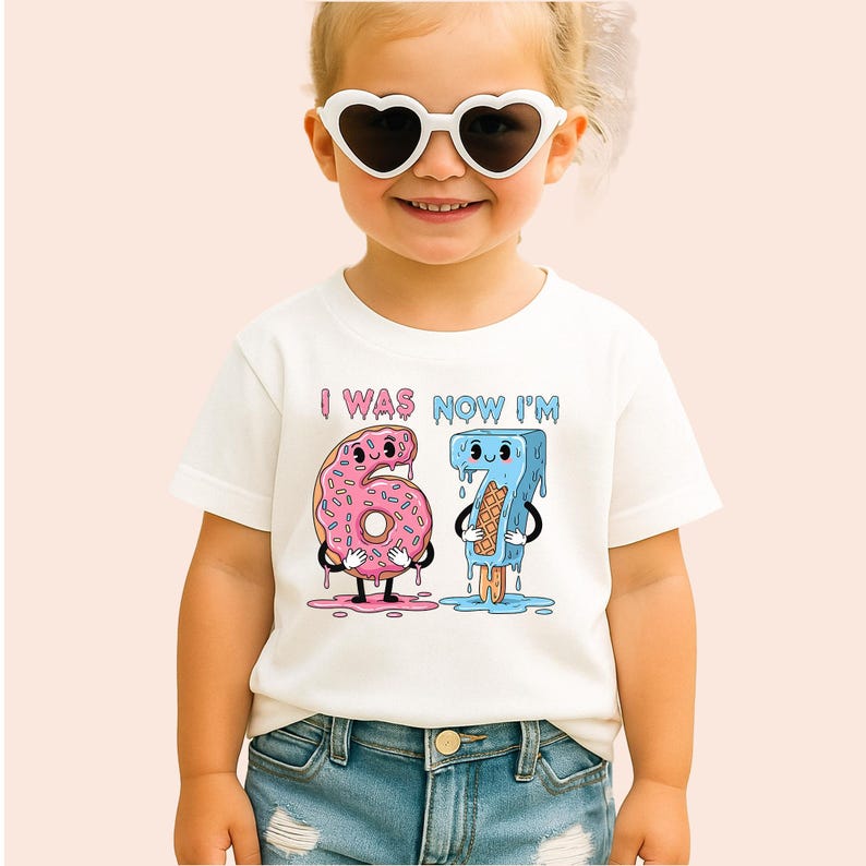 May include: White t-shirt with the text "I WAS NOW I'M" above a cartoon donut and popsicle. The donut is pink with sprinkles and the popsicle is blue. The child is wearing heart-shaped sunglasses.