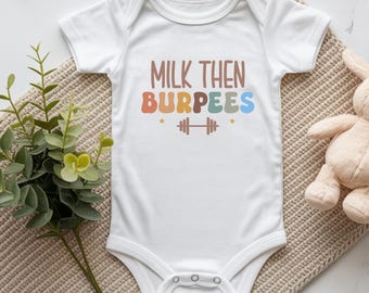 May include: A white short-sleeved baby bodysuit featuring the text MILK THEN BURPEES in multi-colored, rounded lettering. Below the text is a brown barbell icon flanked by two small stars. The garment has a round neckline and three snap closures.