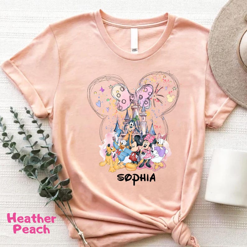 A pink t-shirt with a Disney character design, including Mickey Mouse, Donald Duck, and other iconic figures, along with a castle and fireworks. The text "Sophia" is also visible.