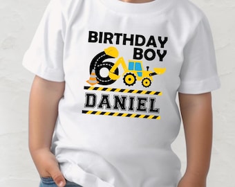 May include: A white t-shirt featuring a graphic with the text BIRTHDAY BOY and the name DANIEL. The design includes a large black number 6 shaped like a road, a yellow construction excavator, a traffic cone, and yellow and black caution stripes.