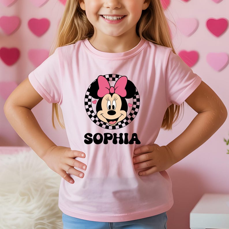 A young girl wearing a pink t-shirt with a Minnie Mouse design and the name "Sophia" printed on it.