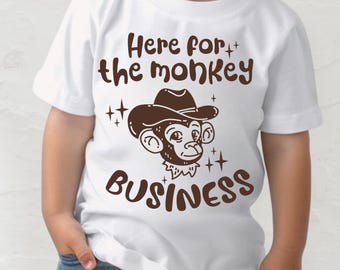 May include: White t-shirt with a brown graphic of a monkey wearing a cowboy hat. The text "Here for the monkey BUSINESS" is printed above and below the monkey illustration. The shirt is worn by a child.