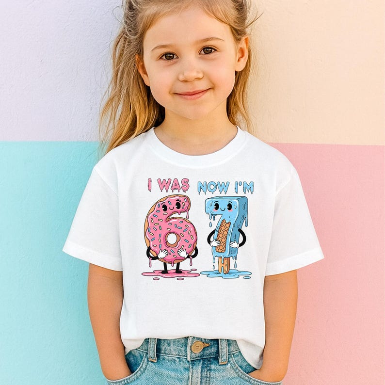 May include: White t-shirt with the text "I WAS NOW I'M" above a cartoon graphic of a pink donut and a blue popsicle. The donut is shaped like the number 6, and the popsicle is shaped like the number 7.