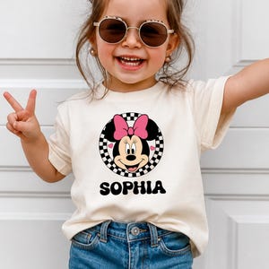 A young girl wearing a white t-shirt with a Minnie Mouse graphic and the name "Sophia" printed on it, smiling and making a peace sign with her hand.
