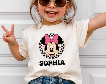 Puede incluir: Una camiseta de manga corta color crema con un gráfico de Minnie Mouse con un lazo rosa dentro de un círculo a cuadros blanco y negro. Debajo del gráfico, el nombre SOPHIA está impreso en letras negras, gruesas y onduladas.