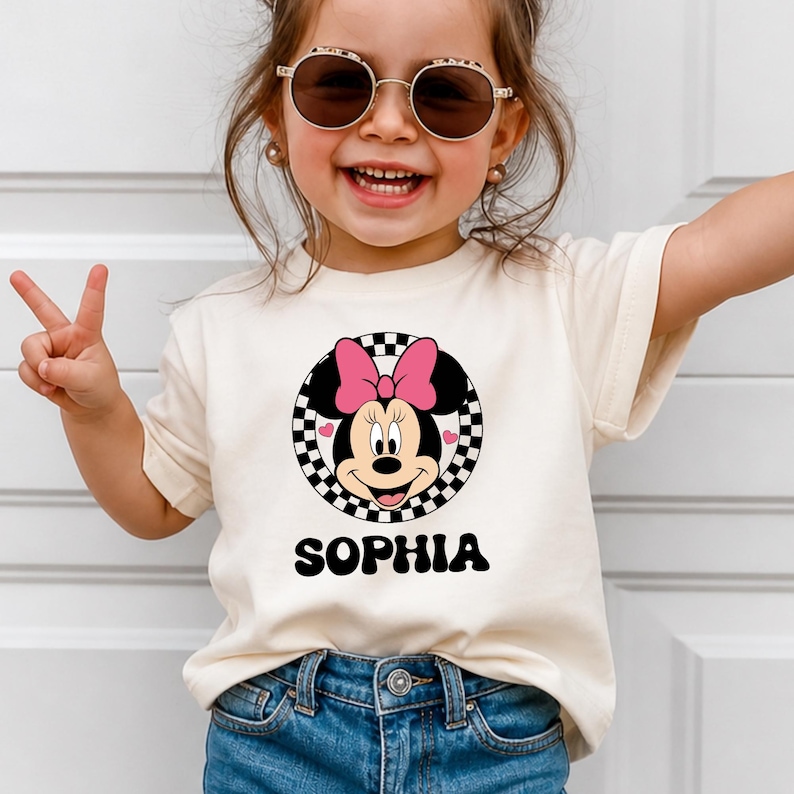 A young girl wearing a white t-shirt with a Minnie Mouse graphic and the name "Sophia" printed on it, smiling and making a peace sign with her hand.