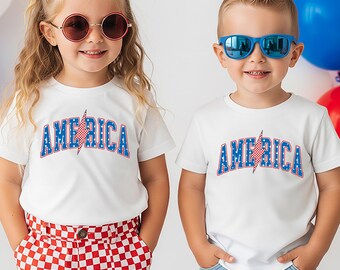Puede incluir: Dos camisetas blancas de manga corta con la palabra AMERICA impresa en azul con estrellas blancas y un rayo a cuadros rojos y blancos que sustituye a la letra I. Las camisetas se llevan con pantalones cortos a cuadros rojos y blancos y vaqueros azules.