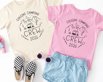 May include: A cream t-shirt and a pink t-shirt, both featuring a black graphic of mountains, a camper, a campfire, and the text COUSINS CAMPING CREW 2026. Also included are light blue denim shorts, a pair of black and white checkered slip-on shoes.