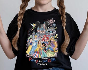 Puede incluir: Una camiseta negra con un gráfico colorido de varias princesas animadas frente a un castillo, un monorraíl y fuegos artificiales. El texto "Party in the USA" está impreso en rojo y azul, seguido de los años "1776-2026" en color blanco.