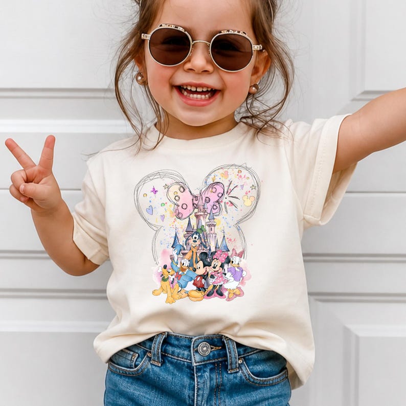 A young girl wearing a white t-shirt with a Disney character design and sunglasses is smiling and making a peace sign.
