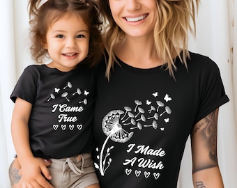 Puede incluir: Camisetas negras a juego con diseños de diente de león blancos. Una camiseta dice "I Came True" con corazones, la otra dice "I Made A Wish" con corazones. Las camisetas las llevan una madre y su hijo.