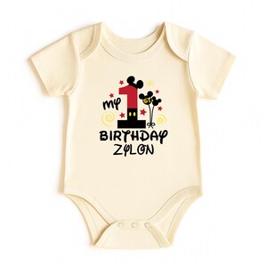 K&ouml;nnte beinhalten: Cremefarbener Baby-Body mit einem Design f&uuml;r den ersten Geburtstag. Das Design beinhaltet die Zahl 1 in Rot und Schwarz, Mickey Mouse Ohren, Sterne und den Text "my BIRTHDAY ZYLON".