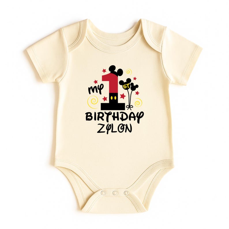 K&ouml;nnte beinhalten: Cremefarbener Baby-Body mit einem Design f&uuml;r den ersten Geburtstag. Das Design beinhaltet die Zahl 1 in Rot und Schwarz, Mickey Mouse Ohren, Sterne und den Text "my BIRTHDAY ZYLON".