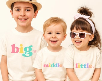 Puede incluir: Tres niños con camisetas crema a juego con las palabras "big", "middle" y "little" en colores pastel. La camiseta "big" tiene un corazón rosa. Un niño lleva un sombrero de paja y otro gafas de sol.