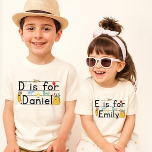 May include: Two off-white short-sleeved t-shirts featuring school-themed graphics. One shirt displays the text 'D is for Daniel' with illustrations of a pencil, apple, paper airplane, and backpack. The other shirt displays 'E is for Emily' with identical icons.